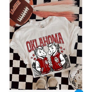 May include: White t-shirt with "OKLAHOMA" in red block letters and two cartoon horse mascots wearing red jackets. The shirt is on a black and white checkered surface with a football and shoes.