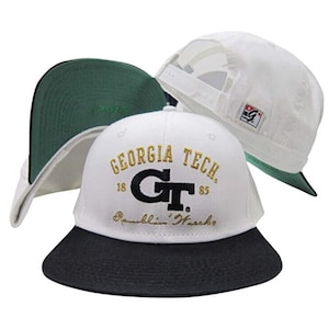 May include: Three white baseball caps with black and green accents. One cap features "GEORGIA TECH" in gold lettering with a black "GT" logo and the words "Ramblin' Wreck". The other two caps have a white crown and a green underside.