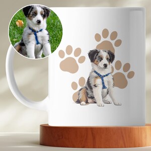 Könnte beinhalten: Weiße Keramiktasse mit einer detaillierten Illustration eines Welpen mit blauem Geschirr, vor einem Hintergrund aus hellbraunen Pfotenabdrücken. Ein kleineres rundes Bild des Welpen ist ebenfalls vorhanden. Die Tasse steht auf einem Holzuntersetzer.