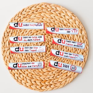May include: Eight silver and red party favors with the word "du" and various celebratory messages in German. The favors are arranged in a circle on a woven straw placemat.