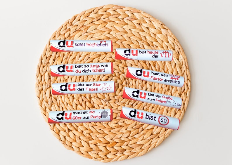 May include: Eight silver and red party favors with the word "du" and various celebratory messages in German. The favors are arranged in a circle on a woven straw placemat.