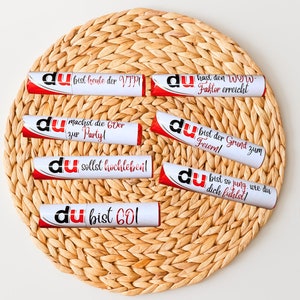 May include: Eight white tubes with red accents and the word "du" printed on them. Each tube has a different message printed on it in German. The tubes are arranged in a circle on a woven straw mat.