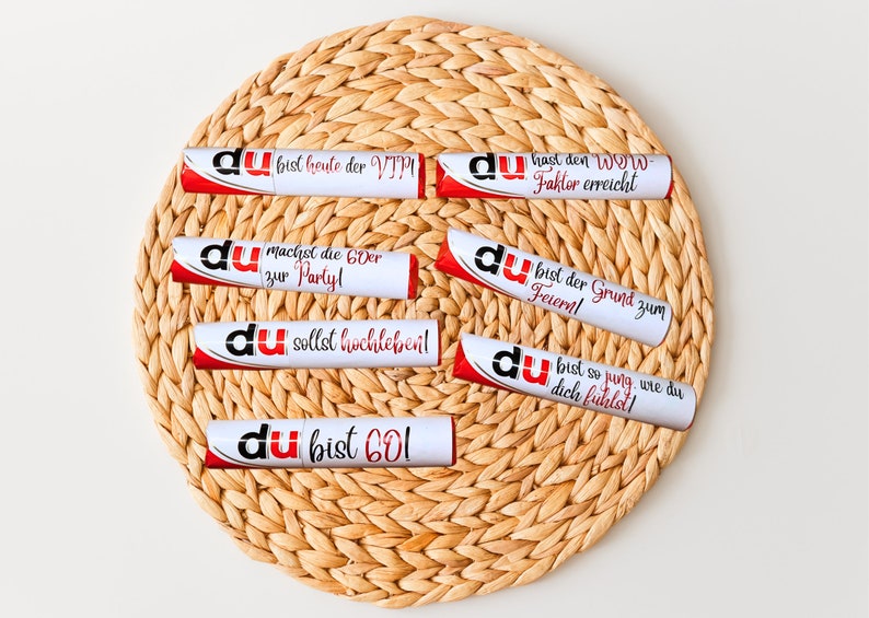 May include: Eight white tubes with red accents and the word "du" printed on them. Each tube has a different message printed on it in German. The tubes are arranged in a circle on a woven straw mat.