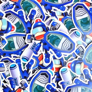 May include: A collection of stickers featuring a blue robot character with a red helmet, visor, and shield. The stickers have a glossy finish and are scattered across the image. The robot has a white arm and a blue weapon.