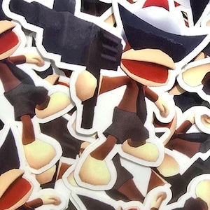 May include: A die-cut sticker featuring a cartoon monkey character wearing a dark brown shirt, dark shorts, and a black hat with a white brim. The character is holding a large, black, blocky rectangular object in one hand. The design is repeated.