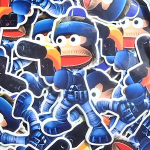 May include: A selection of stickers depicting a cartoon monkey character in a blue uniform, holding a black weapon with an orange handle. The monkey has a blue helmet and a red mouth. The stickers are in different poses.