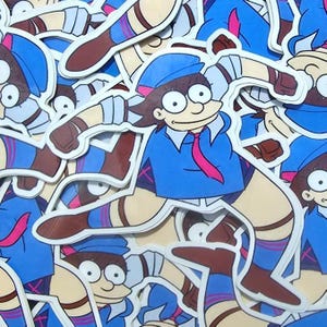 Puede incluir: Un conjunto de pegatinas troqueladas que representan a un personaje de dibujos animados con uniforme azul y corbata rosa. El personaje tiene el pelo castaño y lleva un gorro azul. Las pegatinas tienen bordes blancos y un acabado brillante.