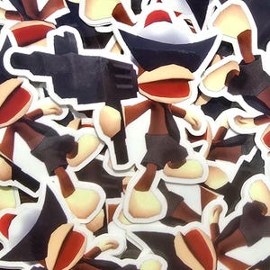 May include: A selection of stickers depicting a cartoon monkey character. The monkey has a brown body, tan hands and feet, and a red mouth. It wears a black hat and shorts, and holds a black gun. The stickers have a white border.