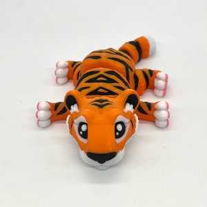 K&ouml;nnte beinhalten: Eine kleine orangefarbene und schwarze Tigerfigur mit wei&szlig;en Akzenten. Der Tiger liegt auf dem Bauch, den Kopf erhoben und die Augen offen.