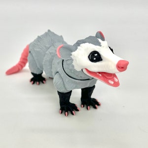 May include: A gray and white 3D printed opossum figurine with pink accents. The opossum has black eyes and a wide open mouth.