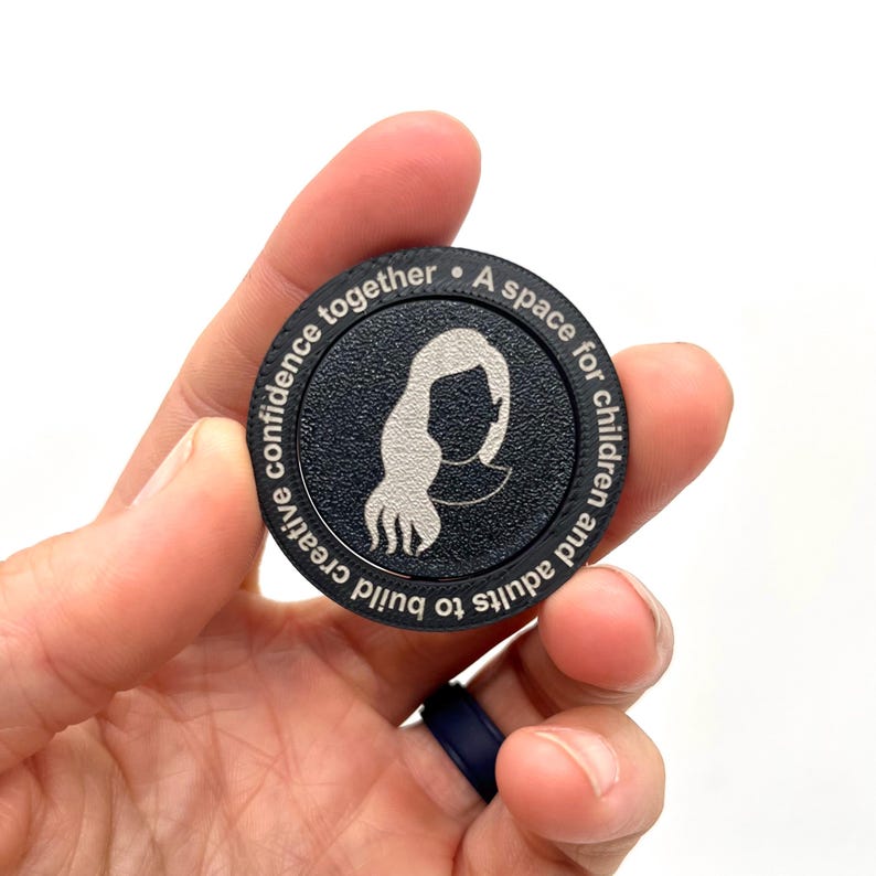 May include: A black round wooden token with a silver silhouette of a person with long hair. The text around the edge reads "A space for children and adults to build creative confidence together."