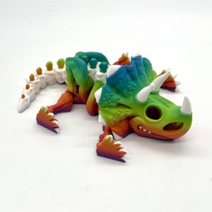 May include: A 3D printed rainbow colored dinosaur toy. The dinosaur is lying on its side with its head turned to the right. The dinosaur has a long tail and three horns on its head.