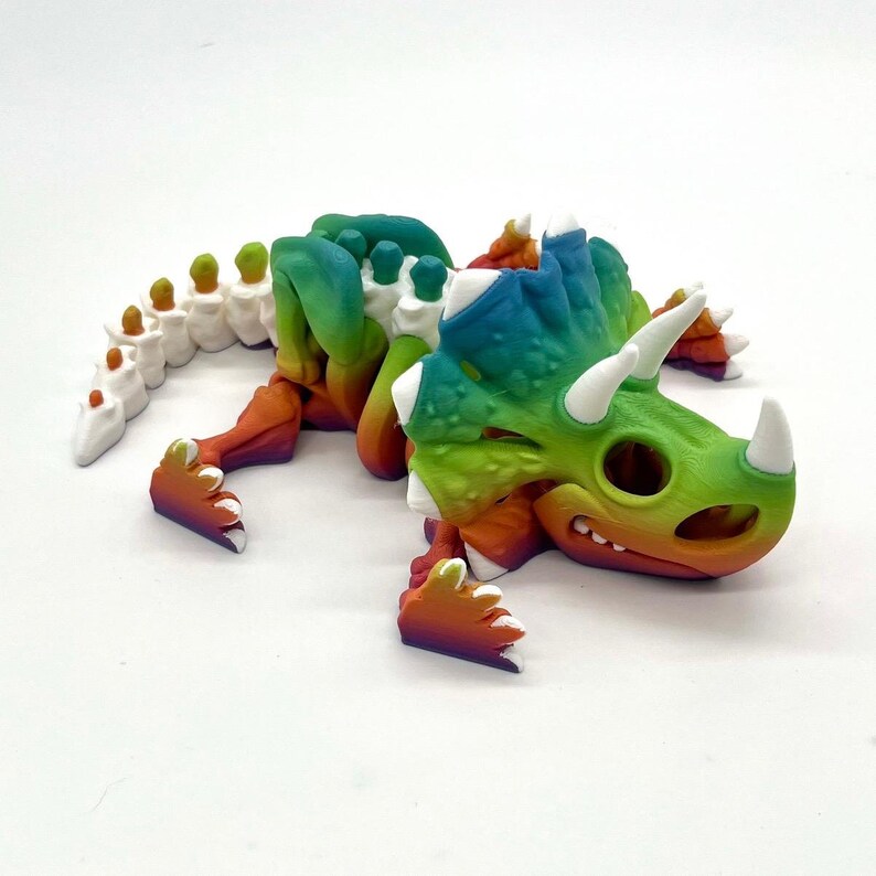 May include: A 3D printed rainbow colored dinosaur toy. The dinosaur is lying on its side with its head turned to the right. The dinosaur has a long tail and three horns on its head.