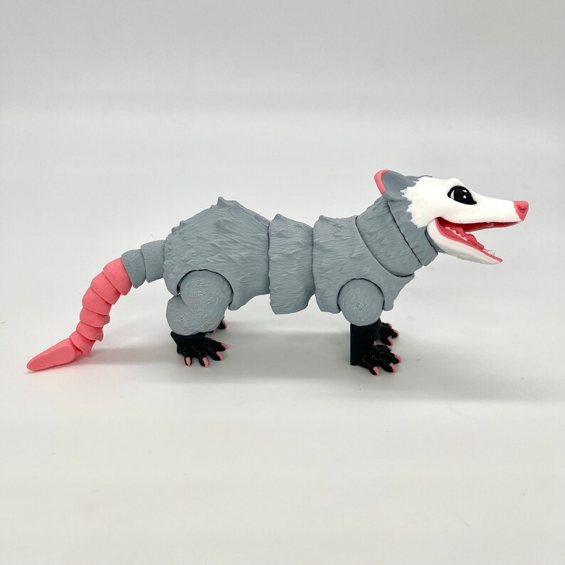 May include: A gray and pink articulated possum toy with a segmented tail and black eyes. The possum is posed standing on all fours with its mouth open.