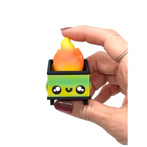 May include: A miniature, whimsical dumpster with a gradient green and yellow body, black wheels, and a smiling face. The dumpster contains a flame-shaped object with orange and yellow hues. The item is held by a hand against a white background.