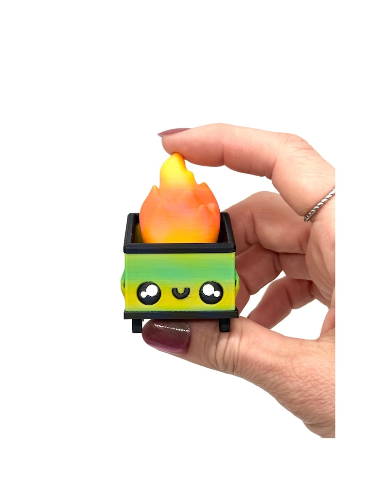 May include: A miniature, whimsical dumpster with a gradient green and yellow body, black wheels, and a smiling face. The dumpster contains a flame-shaped object with orange and yellow hues. The item is held by a hand against a white background.