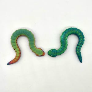 May include: Two green and iridescent segmented plastic toy snakes with a slight bend in their bodies. The snakes are facing each other with their heads touching.