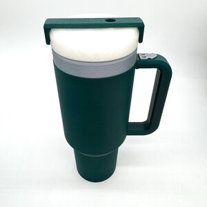 May include: A dark green travel mug with a white interior and a handle. The mug has a gray band around the middle and a dark green lid with a small hole. The mug is designed to keep beverages hot or cold.