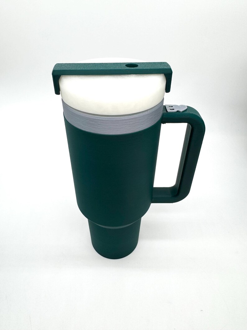 May include: A dark green travel mug with a white interior and a handle. The mug has a gray band around the middle and a dark green lid with a small hole. The mug is designed to keep beverages hot or cold.