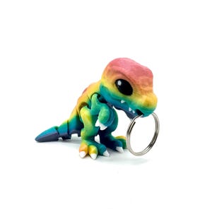May include: Rainbow coloured dinosaur keychain with a silver key ring. The dinosaur is in a crouching position with its mouth open and holding the key ring in its mouth.