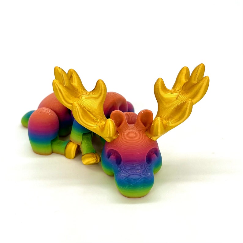 Op de afbeelding: Een regenboogkleurig elandbeeldje met gouden geweien. Het eland ligt met zijn kop op de grond.
