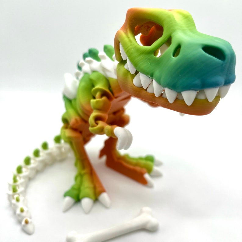 May include: A rainbow colored plastic dinosaur skeleton toy with a white bone. The dinosaur is standing on a white surface.