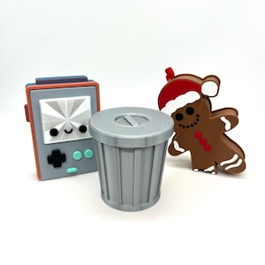 May include: A gray trash can, a gray and red handheld video game console with a happy face, and a brown gingerbread man with a red Santa hat.