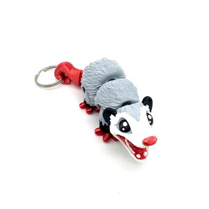 May include: A grey and white possum keychain with red accents. The possum has a curled tail and a toothy grin.