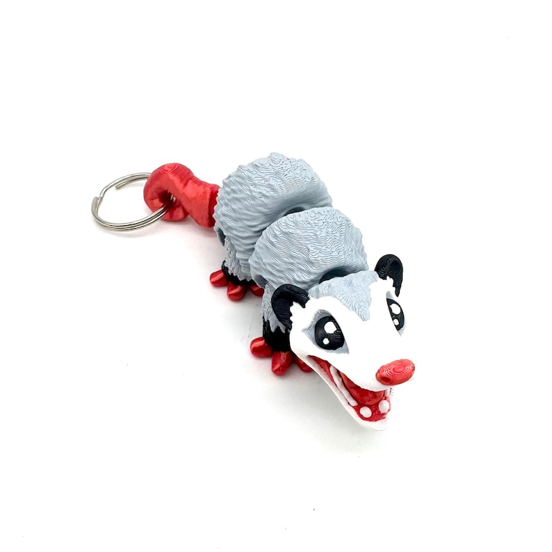 May include: A grey and white possum keychain with red accents. The possum has a curled tail and a toothy grin.
