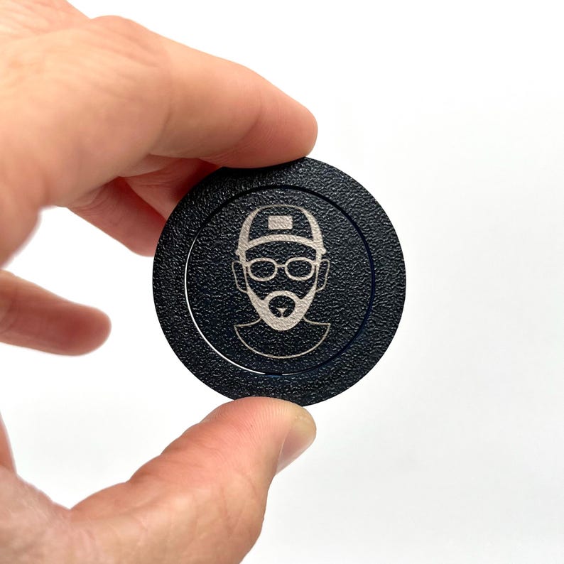 May include: A black poker chip with a white silhouette of a person wearing a cap and glasses.