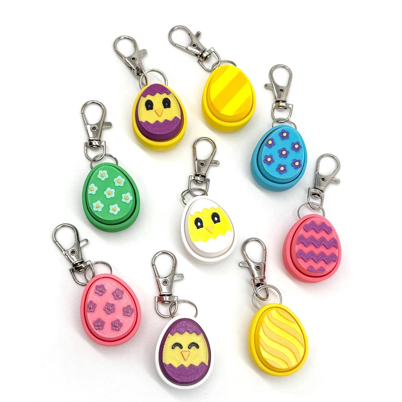 May include: Nine colorful Easter egg-shaped keychains with different designs. Each keychain features a chick, flowers, stripes, or zigzags. The keychains are made of plastic and have a silver metal ring.