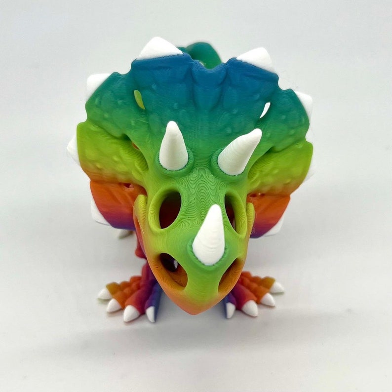 May include: A rainbow-colored 3D printed triceratops dinosaur figurine with white horns and a hollow interior.