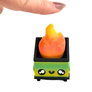 May include: A miniature, 3D-printed dumpster with a smiling face, holding a flame. The dumpster is green and black, and the flame is orange and yellow. The object is on a white background.