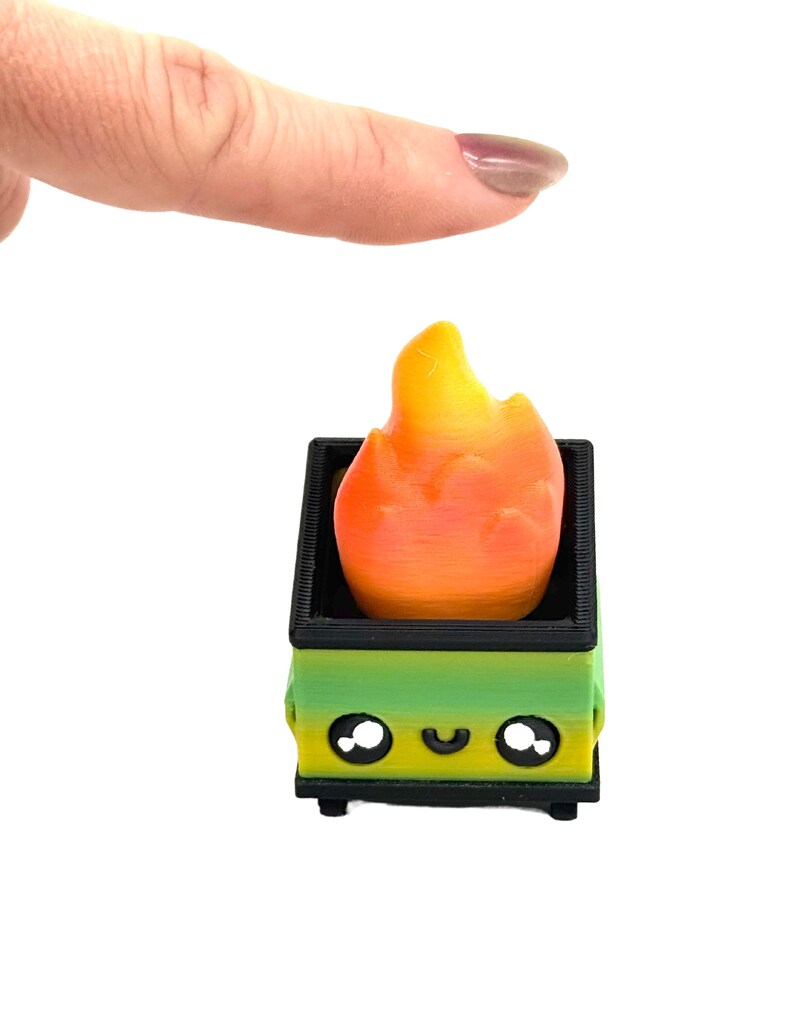 May include: A miniature, 3D-printed dumpster with a smiling face, holding a flame. The dumpster is green and black, and the flame is orange and yellow. The object is on a white background.
