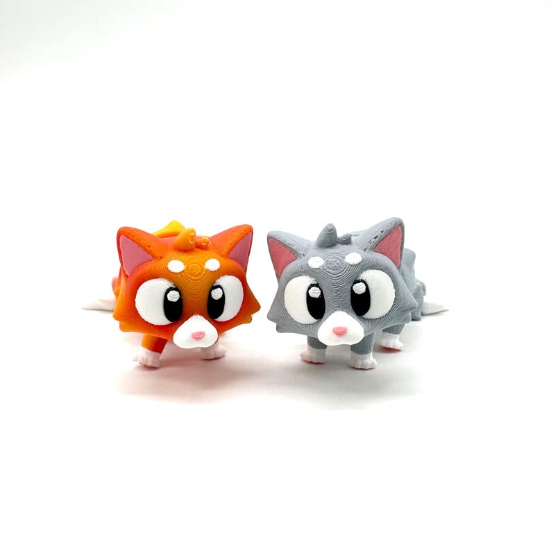May include: Two small, 3D printed cat figurines. One is orange and the other is grey. Both have large, white eyes and pink noses.
