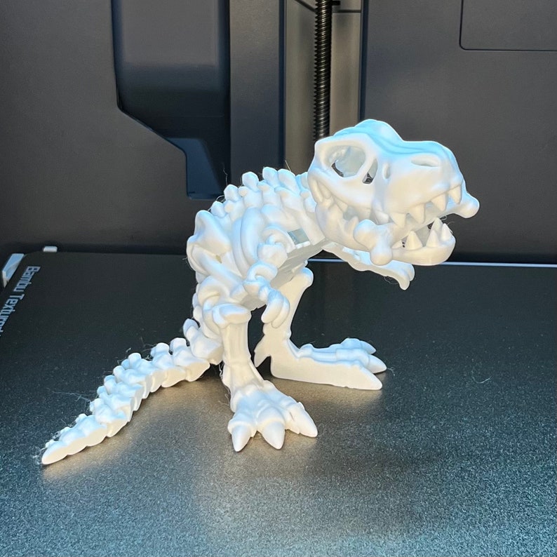 May include: A white plastic dinosaur skeleton model. The dinosaur is posed in a standing position with its mouth open. The model is detailed with visible ribs, vertebrae, and leg bones.