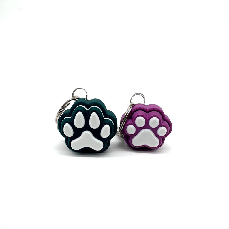 May include: Two paw print shaped keychains, one is teal green and the other is purple. Both have a white paw print design.