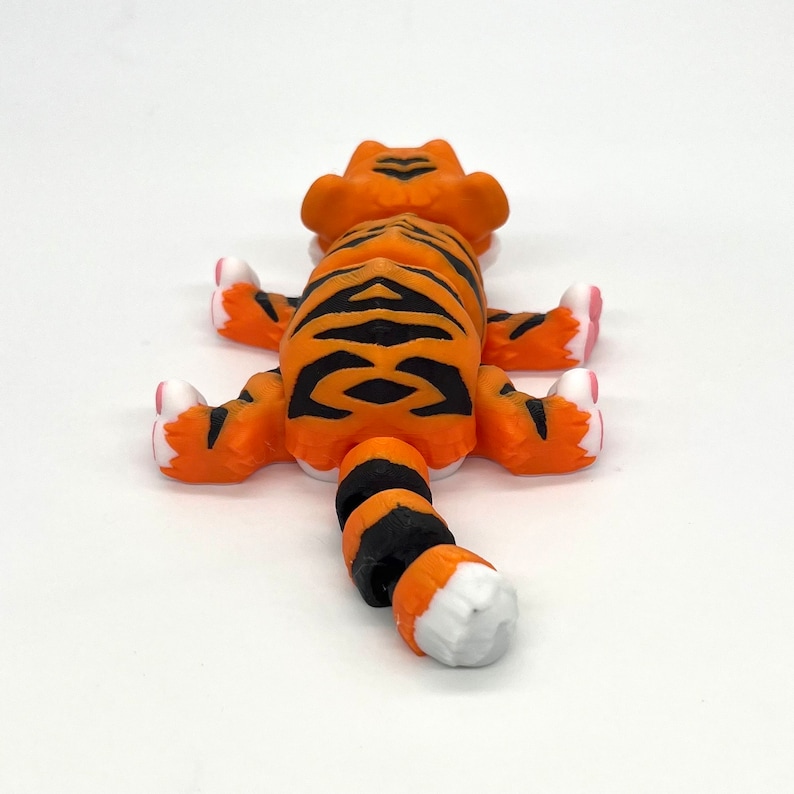 K&ouml;nnte beinhalten: Orangefarbene und schwarze Tigerfigur mit wei&szlig;en Akzenten. Der Tiger liegt auf dem Bauch, die Beine ausgestreckt und der Schwanz eingerollt.