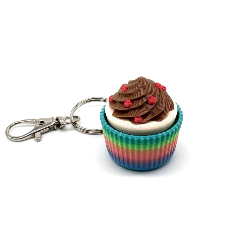 May include: A rainbow cupcake keychain with a chocolate frosting and red sprinkles. The keychain has a silver metal ring and a silver clip.