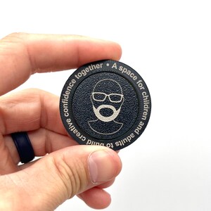 May include: A black round token with a white outline and text that reads "A space for children and adults to build creative confidence together."  The token features a white silhouette of a person wearing glasses and a beard.