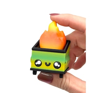 May include: A 3D-printed miniature dumpster with a smiling face and a flame on top. The dumpster is green and yellow with black accents and wheels. The flame is orange and yellow.