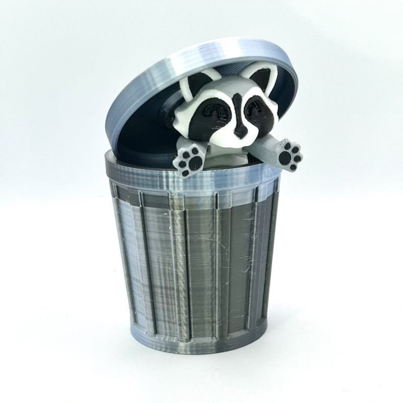 May include: A gray and black trash can with a raccoon figurine inside. The raccoon is peeking out of the top of the can with its paws on the rim.