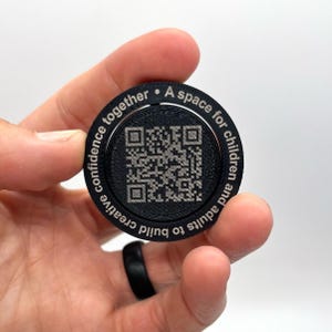 May include: Black round disc with a QR code in the center and the text "A space for children and adults to build creative confidence together." around the edge.