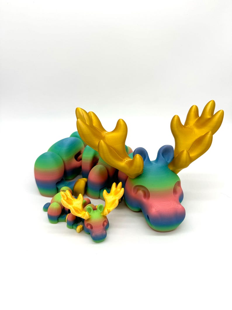 Op de afbeelding: Drie regenboogkleurige elandfiguren met gouden geweien. De grootste eland ligt met zijn geweien omhoog. De twee kleinere elanden liggen naast elkaar.