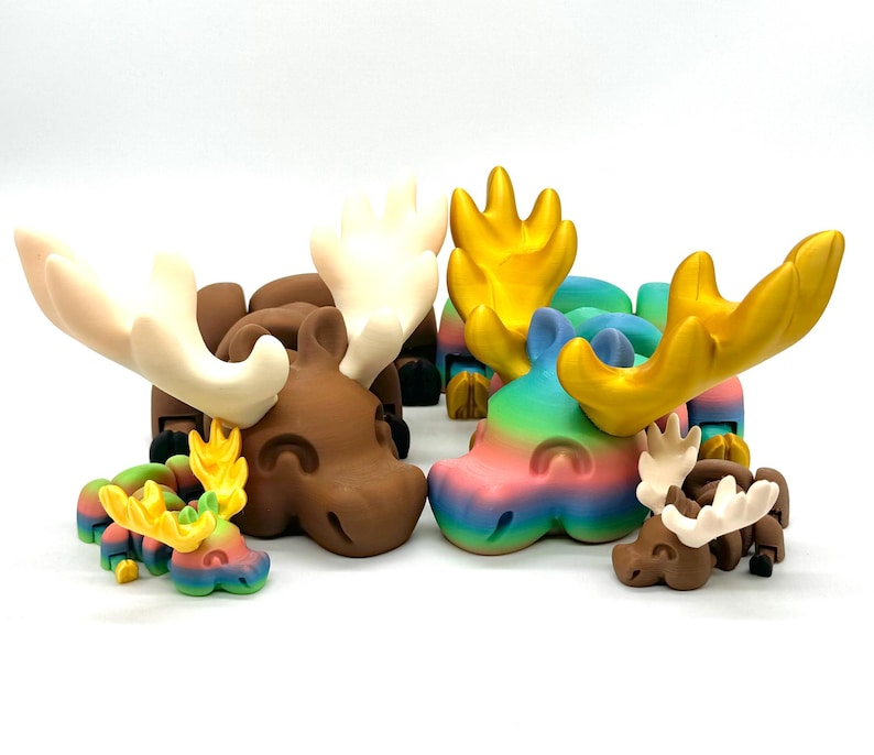 Op de afbeelding: Een set van vijf eland-beeldjes, elk met een ander kleurenschema. De elanden zijn gemaakt van een glad, plastic-achtig materiaal. De kleuren omvatten bruin, wit, goud en een regenboogkleurige gradi&euml;nt.