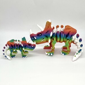 May include: Two rainbow colored dinosaur skeletons, one larger than the other, are posed on a white surface. The larger dinosaur has three horns on its head and a long tail. The smaller dinosaur has two horns on its head and a shorter tail.