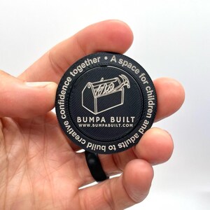 May include: A black round token with silver lettering. The token features a toolbox with tools inside and the text "BUMPA BUILT" with the website address "WWW.BUMPABUILT.COM". The text around the edge of the token reads "A space for children and adults to build creative confidence together."