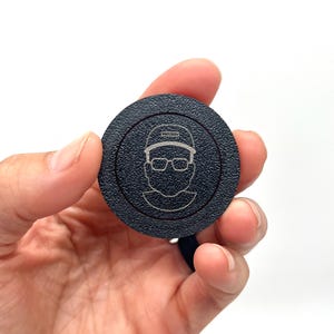 May include: A black round disc with a white outline and a white line drawing of a person wearing a hat and glasses. The person's hat has the word "PROUD" written on it.