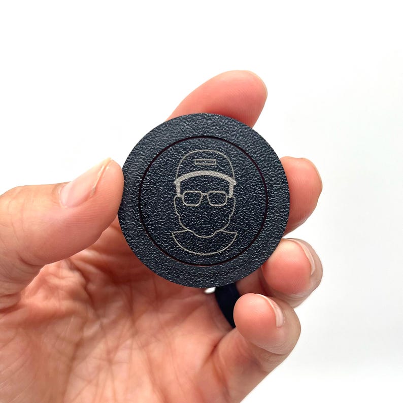 May include: A black round disc with a white outline and a white line drawing of a person wearing a hat and glasses. The person's hat has the word "PROUD" written on it.