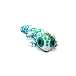 May include: White gecko keyring with green spots and blue eyes. The gecko is holding a silver key ring.